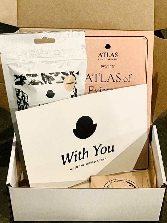 An open Comfort Box holding a With You card, a copy of The Atlas of Existence, a pouch of Atlas tea, and an engraved wooden coaster.