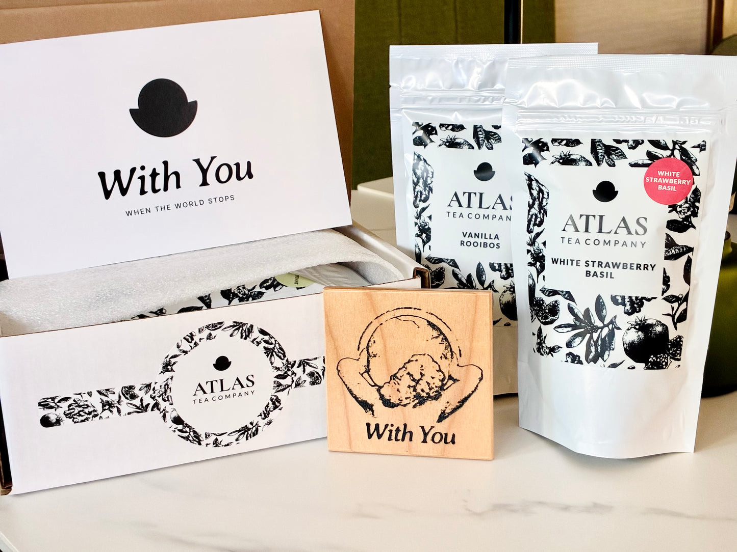 Comfort Box contents laid out: the With You card, the illustrated box sleeve, an engraved wooden coaster, and two pouches of Atlas tea.